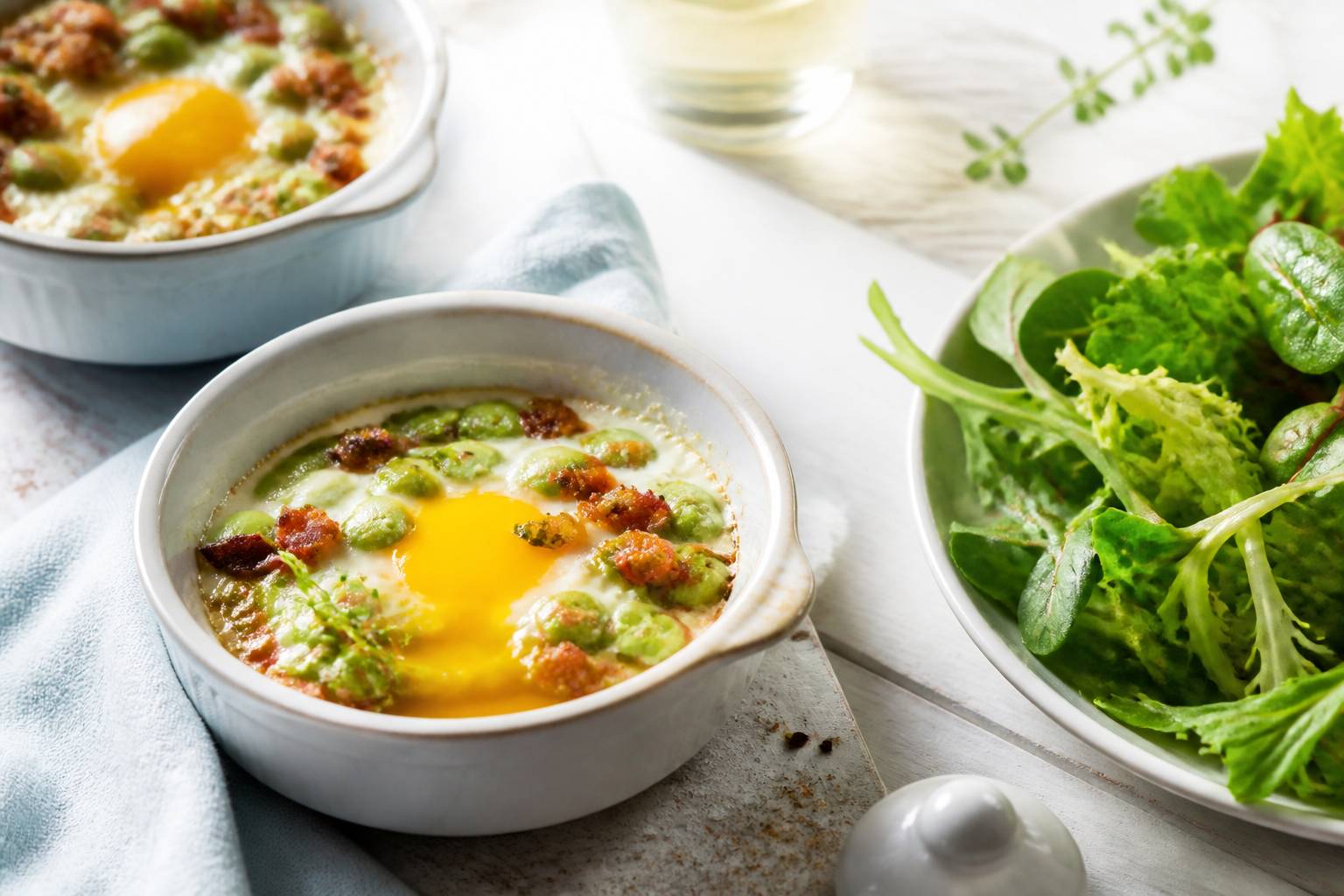 Ramequins d'œufs cocotte dorés aux fèves vertes et lardons fumés avec jaune coulant sur table claire